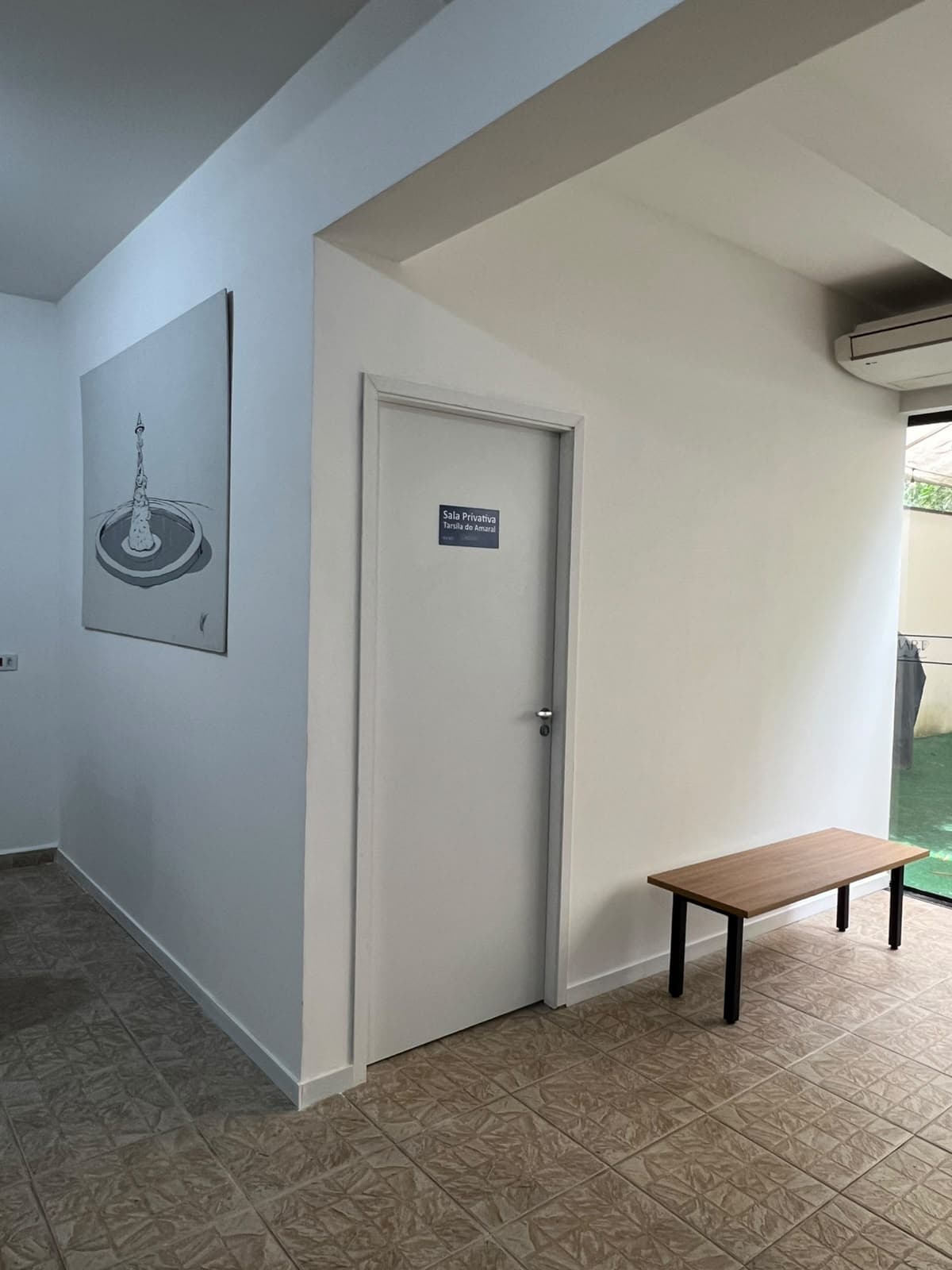 Acesso à sala privativa Tarsila do Amaral, com a placa de identificação, banco e jardim ao fundo